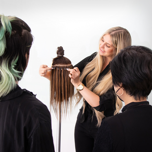 Two people watching a person demonstrate hair extension application